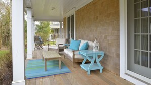 Une véranda couverte avec un plancher en bois, une colonne blanche, un tapis rayé bleu et vert, une table en bois, un canapé blanc avec des coussins bleus et une petite table d’appoint bleue surmontée d’une lanterne.