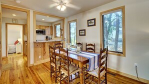 Uma sala de jantar com mesa de madeira, cadeiras e um ventilador de teto. Cozinha visível ao fundo, com armários de madeira e micro-ondas.