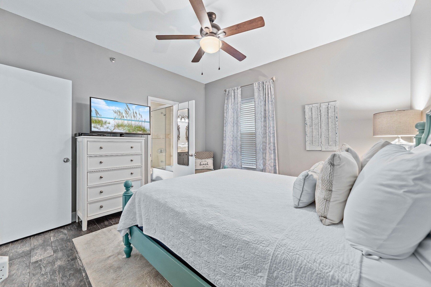 Une chambre à coucher comprenant un lit, une commode, un téléviseur, un ventilateur de plafond et une fenêtre avec des rideaux.