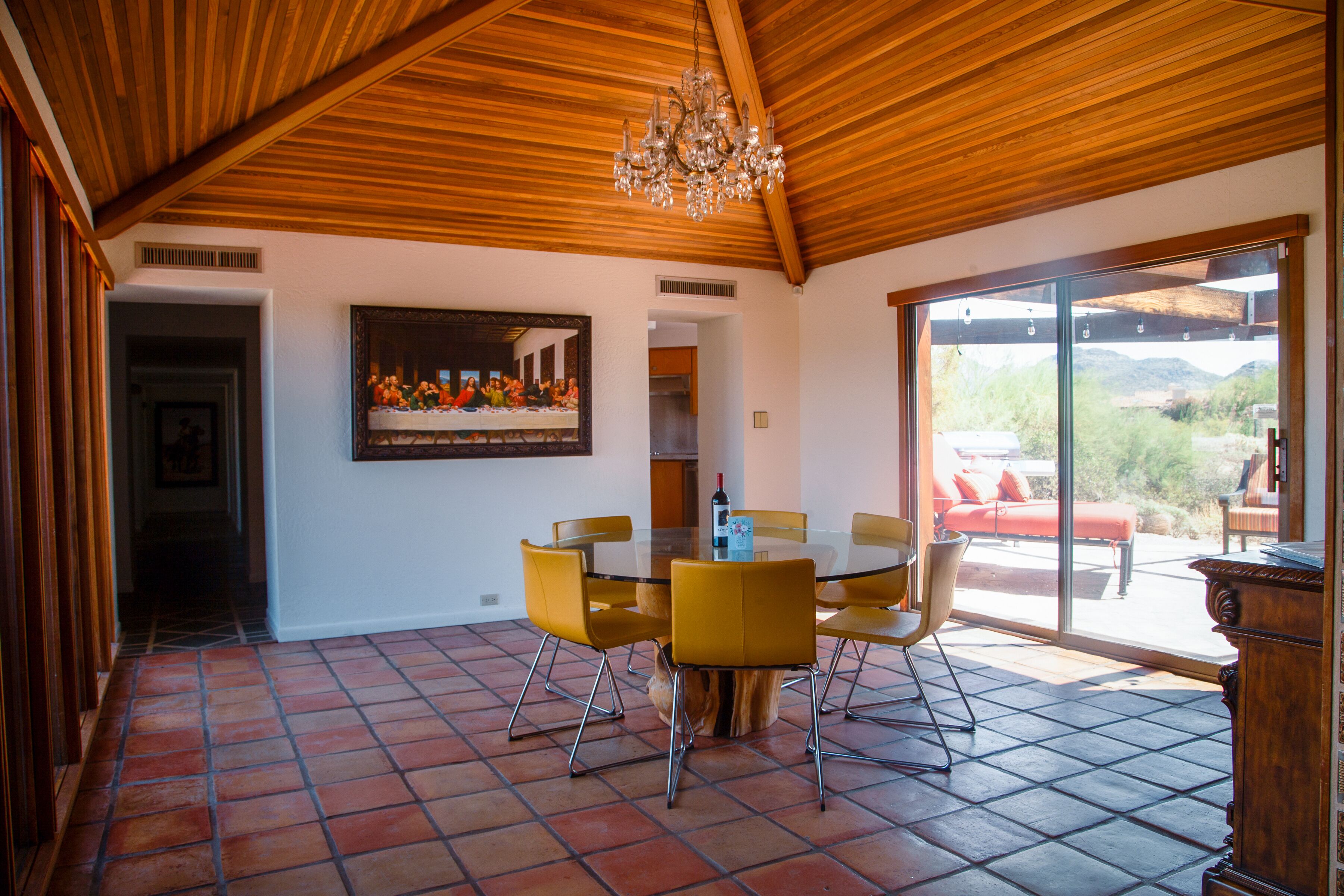 Een eetruimte met een glazen tafel en gele stoelen, een houten plafond en een groot schilderij van een religieuze scène.