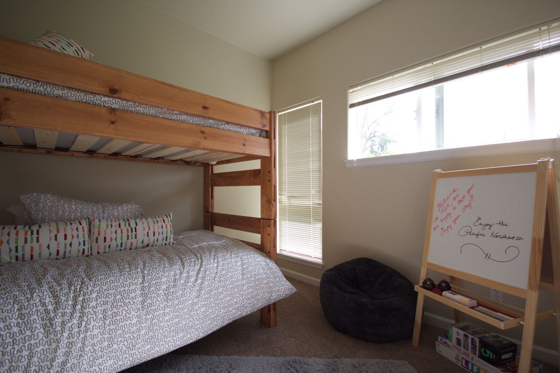 A bunk bed with patterned bedding, a whiteboard with writing, and a bean bag chair in a room with a large window.