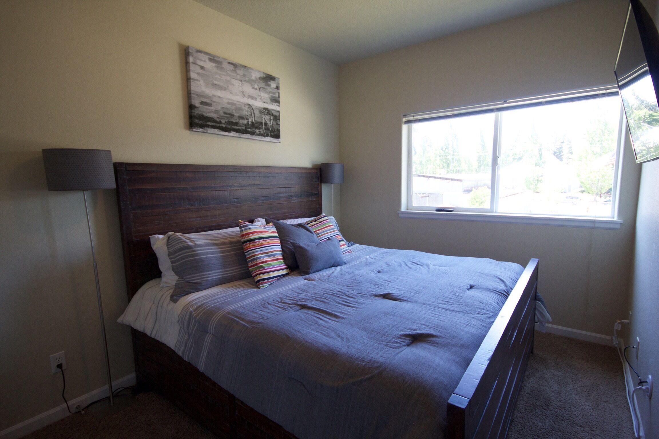 A bedroom with a bed, a nightstand, a lamp, a window, and a wall-mounted artwork.