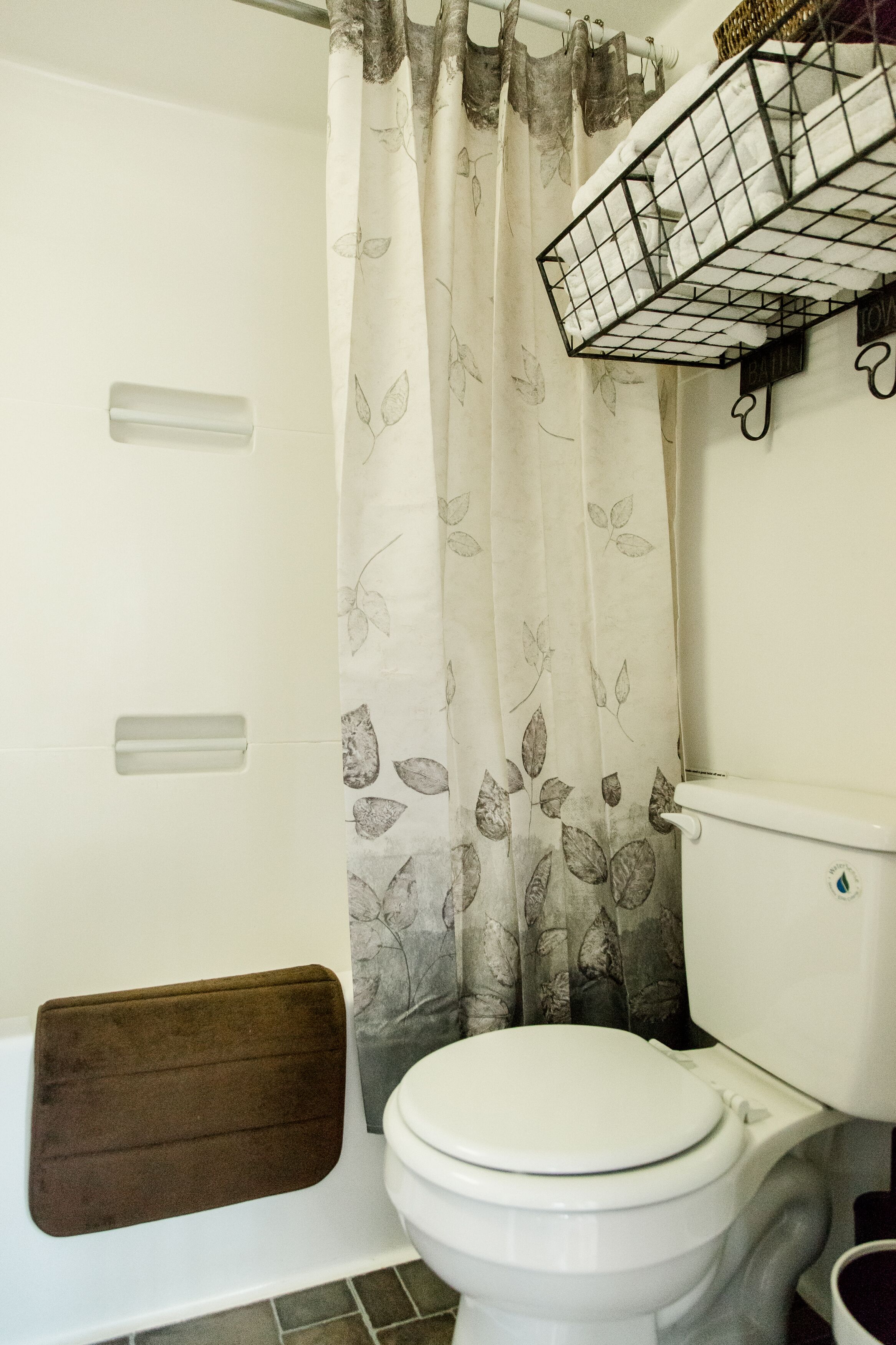 Un baño con inodoro, cortina de ducha estampada con hojas, toallero y bañera con un tapete marrón.