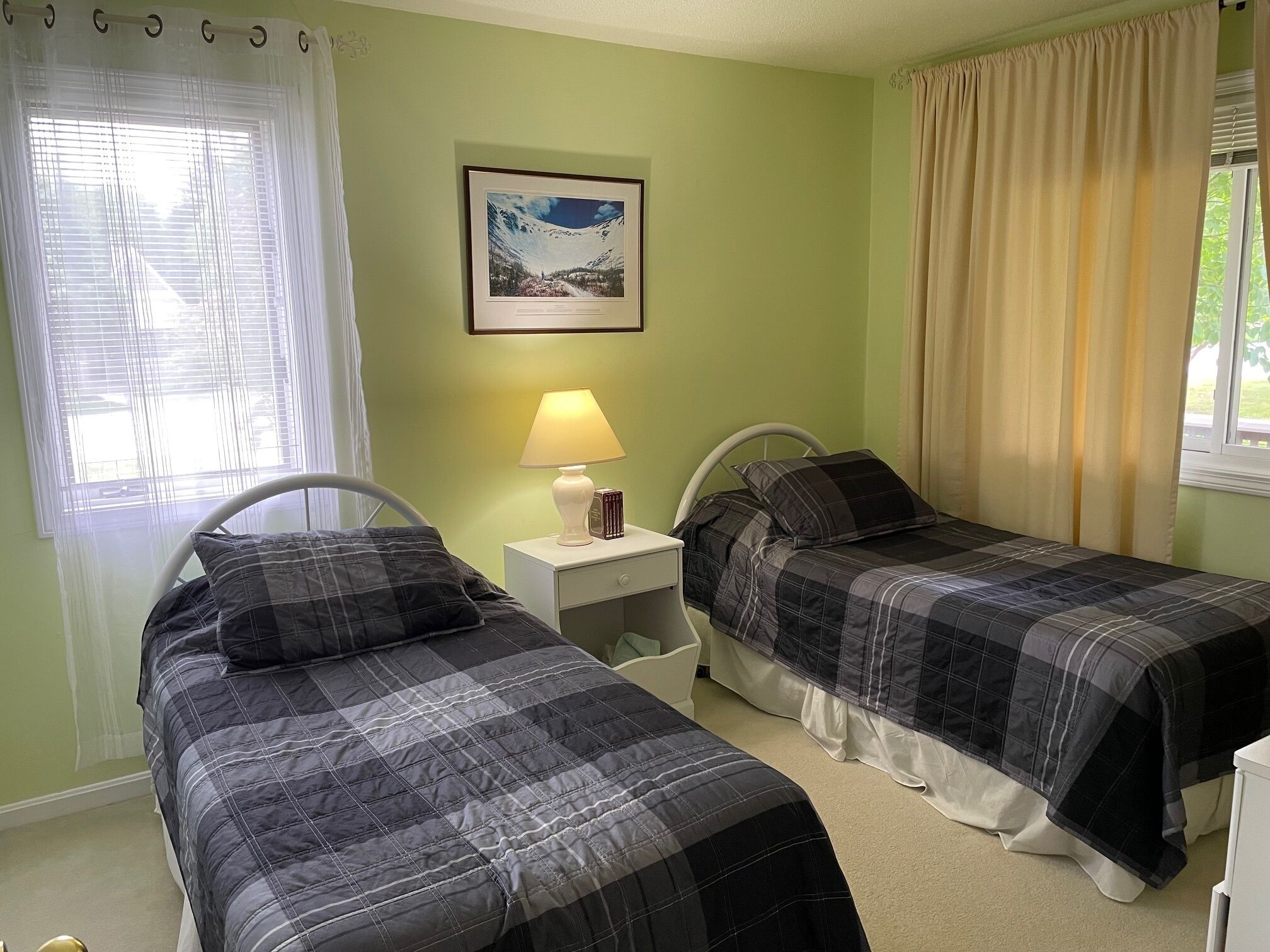 A room with two beds, a nightstand with a lamp, a framed picture on the wall, and a window with curtains.