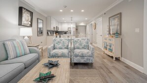 A modern living room with a grey sofa, two patterned armchairs, a coffee table, and a kitchen in the background. - Downtown Dewey Beach Ocean/Bay Views 2BR Pools Fire Pit (Rehoboth Beach)