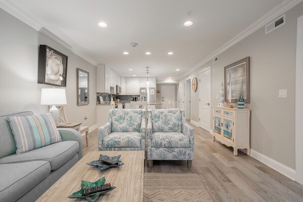 A modern living room with a grey sofa, two patterned armchairs, a coffee table, and a kitchen in the background.