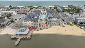 An aerial view of a coastal town with a marina, a beach, and a large hotel with multiple floors and balconies. - Downtown Dewey Beach Ocean/Bay Views 2BR Pools Fire Pit (Rehoboth Beach)