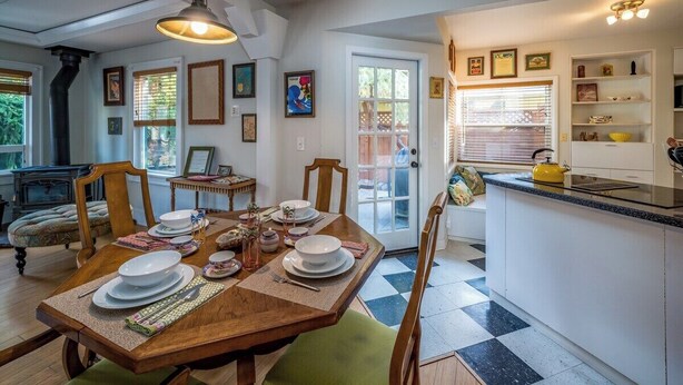 Un comedor con una mesa de madera dispuesta para cuatro personas, rodeada de sillas. Se observa una zona de cocina con una encimera y un hervidor amarillo.