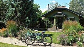 Una pequeña casa verde con chimenea, rodeada por un jardín con diversas plantas y flores. Dos bicicletas están estacionadas frente a la casa.