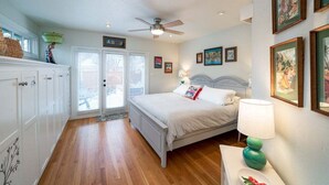 Un dormitorio con cama, ventilador de techo, mesita de noche con lámpara y ventana con persianas.