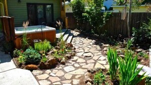 Un patio trasero con jacuzzi, camino de piedra y diversas plantas.