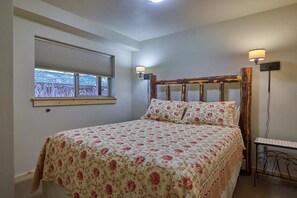 Ein Schlafzimmer mit einem Bett, einem Fenster mit Jalousien, einer Wandlampe und einem kleinen Tisch.