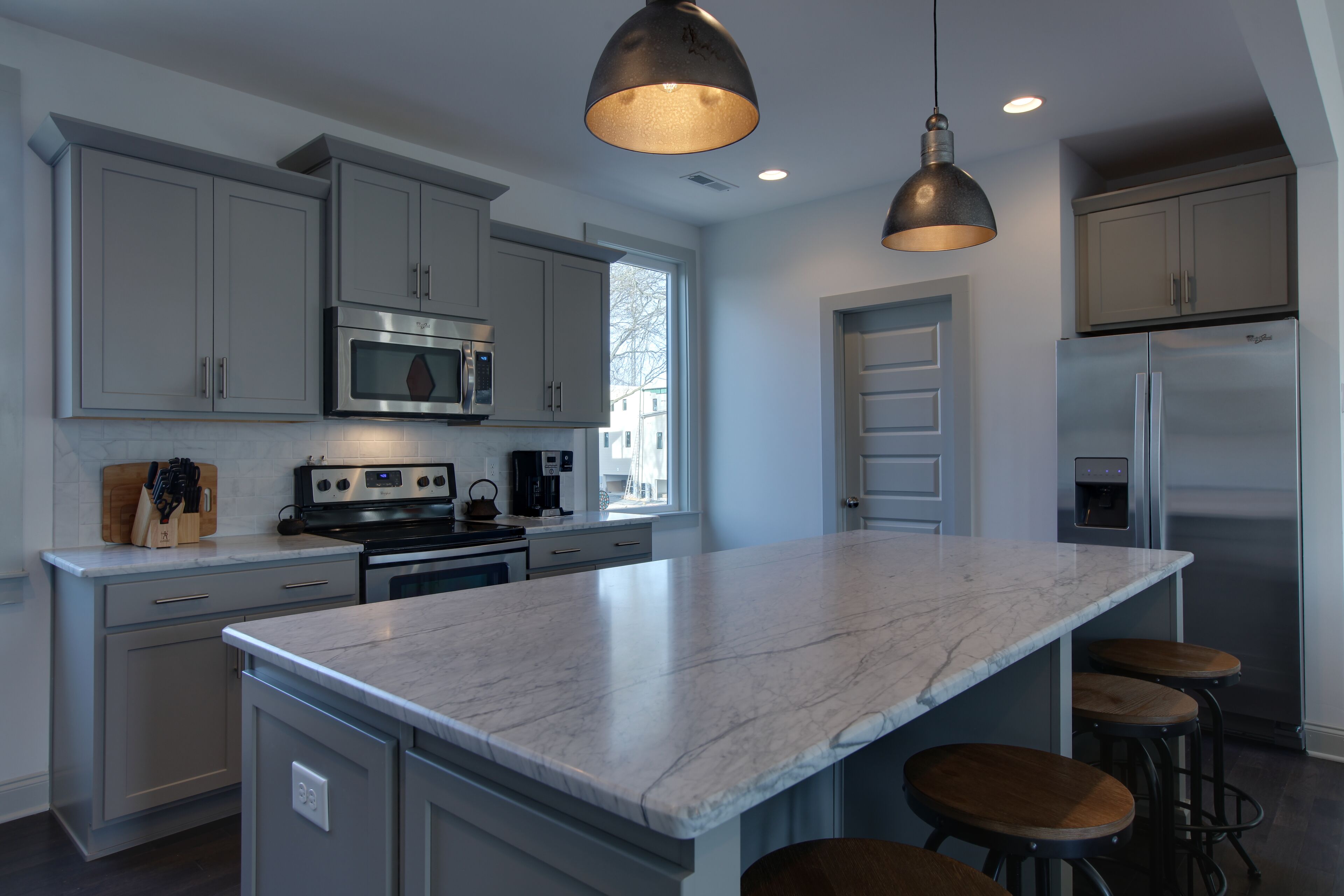 A modern kitchen with grey cabinets, a large marble island, stainless steel appliances, and pendant lighting.