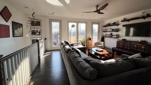 A modern living room with a large sofa, a coffee table, and a TV. There is a staircase with a railing leading to a balcony. - Music Centric Home, 5 minutes to Broadway. (Nashville)