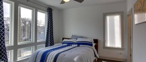Une chambre avec un lit, un ventilateur de plafond, de grandes fenêtres et un tapis blanc.