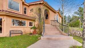 A two-story house with a stone staircase leading to the entrance, surrounded by a well-maintained lawn and trees. - 2 KING Masters, Pikes Peak Views.  Sleeps 17, Close To AF Academy - 2 Living Rms (Colorado Springs)