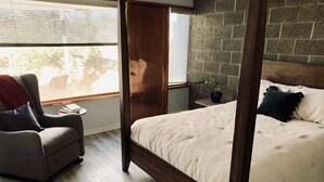 A bedroom with a four-poster bed, a grey armchair, a small table, and a window with blinds. - Architectural dream guest house (Phoenix)