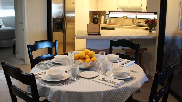 Un coin repas avec une table ronde dressée pour quatre personnes, agrémentée d’assiettes, de bols, de verres et de couverts blancs. Un saladier rempli de citrons est posé sur la table, et une cuisine moderne se trouve au fond de la pièce.