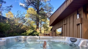 Un bain à remous situé dans un cadre naturel, entouré d’arbres, avec un bâtiment en bois en arrière-plan.
