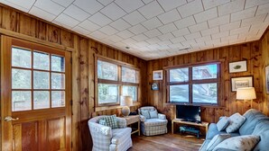 Una habitación acogedora con paredes de madera, ventanas amplias, un sofá, dos sillones, un televisor y una lámpara.