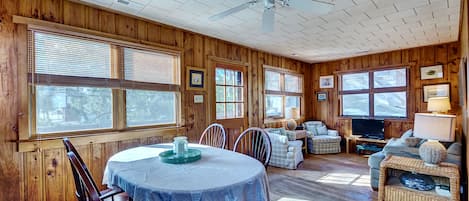 A wooden cabin interior with a dining table, chairs, a sofa, and a television.