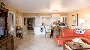 A spacious living room with a red leather sofa, a dining area with wicker chairs, and a kitchen in the background. - SPECTACULAR BEACH CONDO. 2/2 PLUS DEN. HEATED POOL - EMAIL US. (Indian Shores)