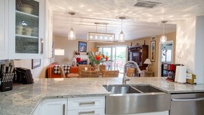 A modern kitchen with a stainless steel sink, white cabinetry, and a dining area with wicker furniture and a vase of flowers. - SPECTACULAR BEACH CONDO. 2/2 PLUS DEN. HEATED POOL - EMAIL US. (Indian Shores)