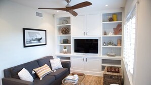 A modern living room with a dark grey sofa, a flat-screen TV, white built-in shelving, and a ceiling fan. - Playa One Bedroom (251) (Carpinteria)