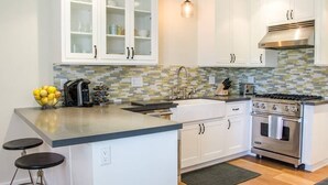 A modern kitchen with white cabinets, a tiled backsplash, a stainless steel stove, and a small island with stools. - Playa One Bedroom (251) (Carpinteria)