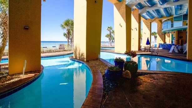 A pool area with a view of the ocean, palm trees, and a covered seating area with tables and chairs. - Corner Condo - Stunning Gulf Views -Comp. Beach Service, Water Park & Lazy River (Panama City Beach)