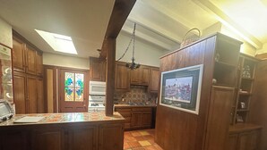 Una cocina con armarios de madera, suelo de baldosas, horno empotrado y un amplio ventanal con puerta de vidrio emplomado.