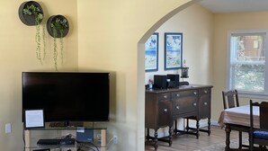 A living room with a flat-screen TV, a glass coffee table, a wooden dining table with chairs, a dark wooden dresser, and two wall-mounted planters with trailing plants. - Manzanita Beach Charmer! Dog Friendly Home with a Short Walk to Beach ~MCA#1012 (Manzanita)