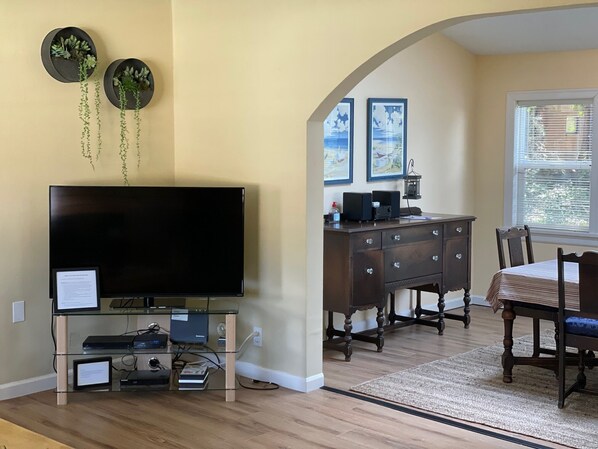 Una sala de estar con televisor de pantalla plana, una mesa de centro de vidrio, una mesa de comedor de madera con sillas, un aparador de madera oscura y dos macetas colgantes en la pared con plantas de cascada.