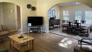 A living room with a TV, dining table, and chairs, a wooden coffee table, and a wicker sofa. - Manzanita Beach Charmer! Dog Friendly Home with a Short Walk to Beach ~MCA#1012 (Manzanita)