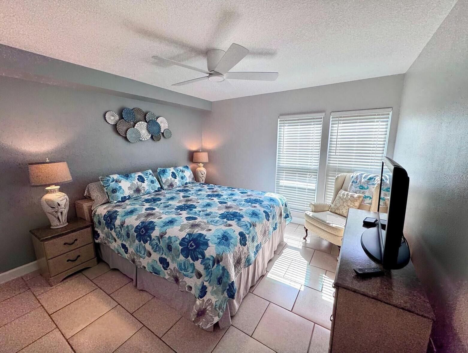 A bedroom with a bed, two bedside tables, a ceiling fan, decorative wall art, a window with blinds, and a television on a stand.