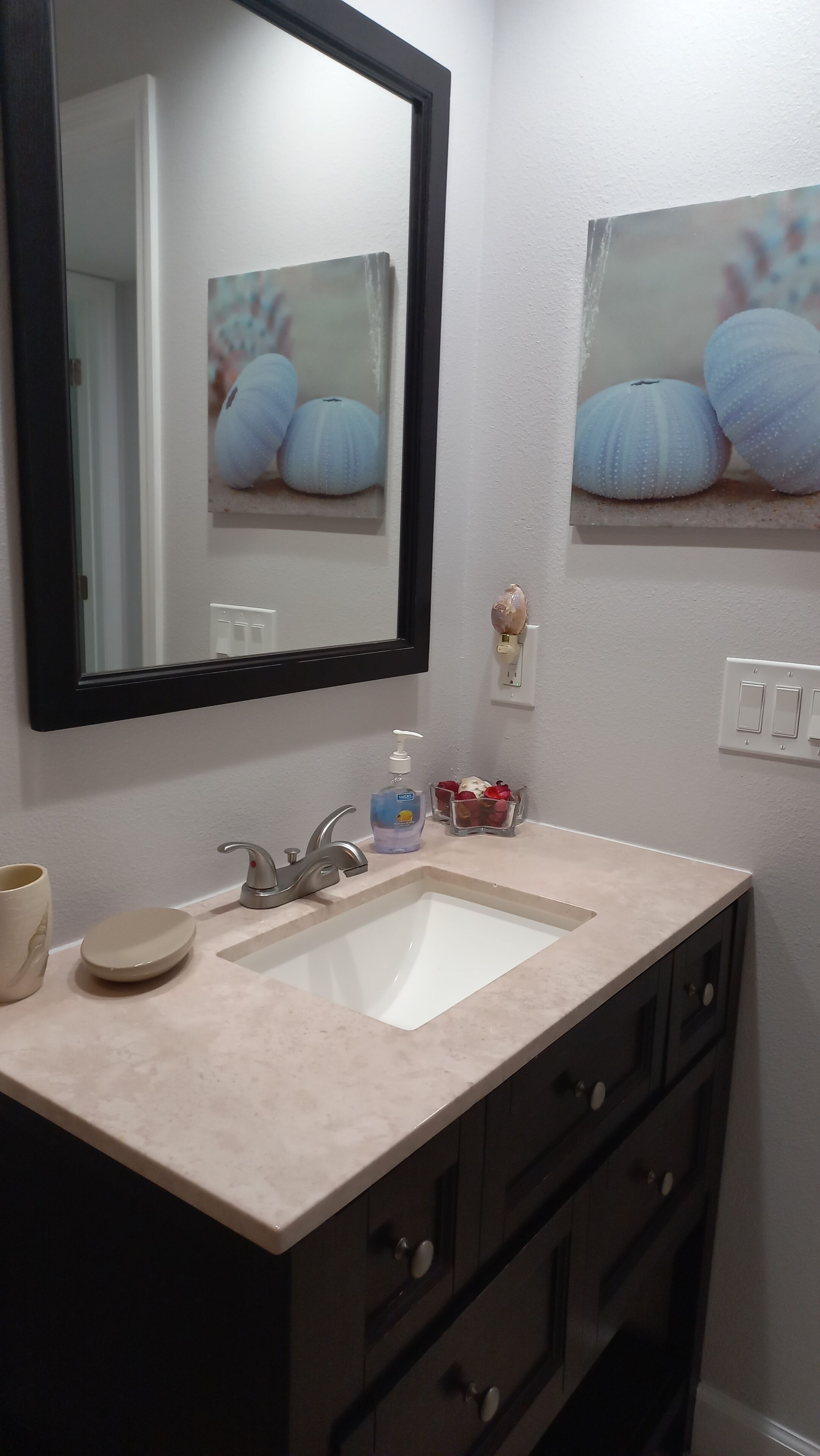 Une salle de bain avec un lavabo blanc, des meubles sombres, un miroir et un interrupteur fixé au mur.