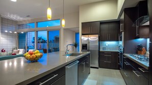 A modern kitchen with stainless steel appliances, a large island with a fruit bowl, and a view of the outdoors through the windows. - PS Paradise: Contemporary Modern Home; Large with Modern Comforts (Palm Springs)