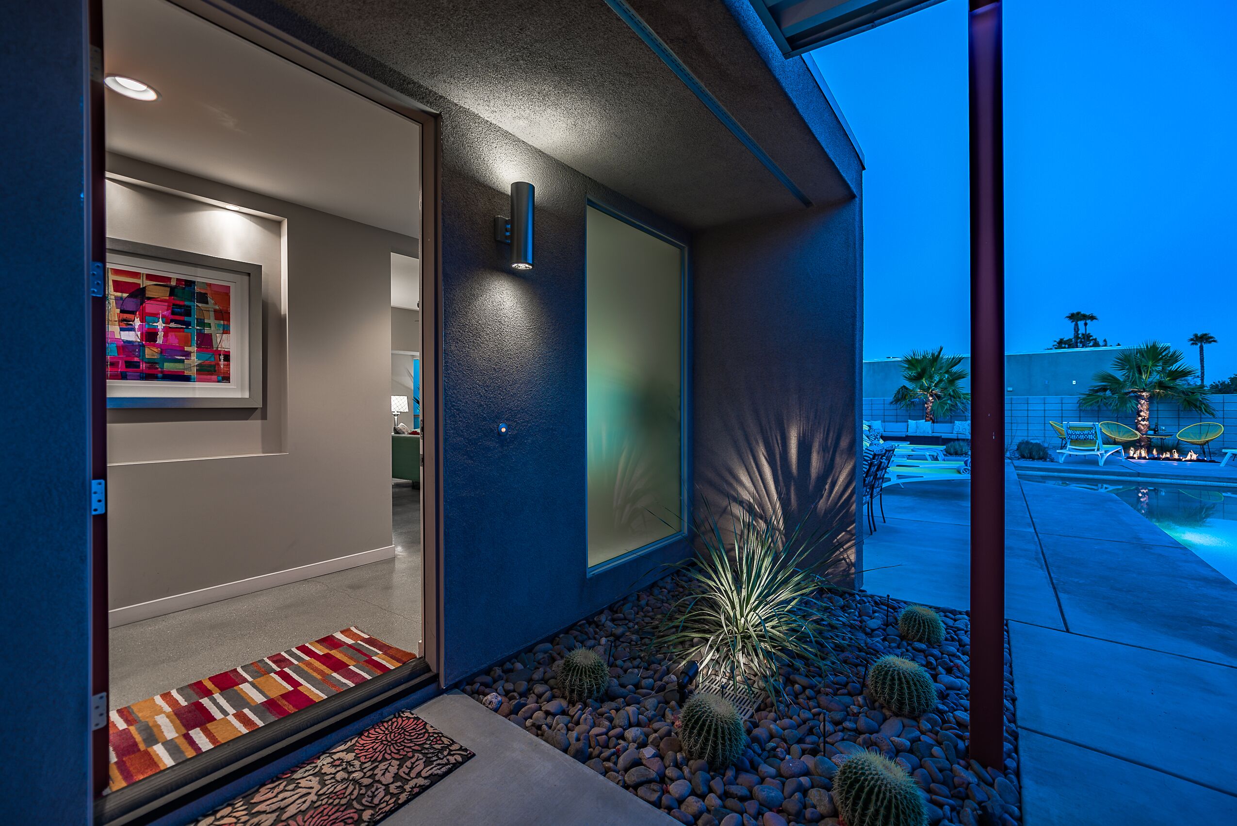 Una entrada de edificio moderno con un colorido tapete, un jardín de cactus y una zona de piscina visible a través de las puertas de cristal.