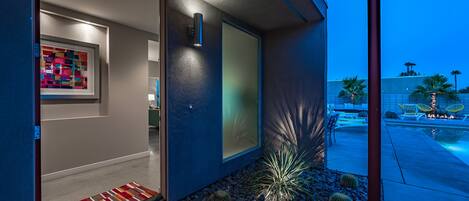 Una entrada de edificio moderno con un colorido tapete, un jardín de cactus y una zona de piscina visible a través de las puertas de cristal.