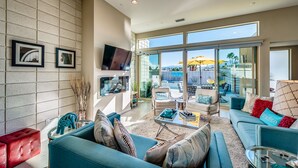 A spacious living room with a large window, a flat-screen TV mounted on the wall, and a red ottoman. - PS Paradise: Contemporary Modern Home; Large with Modern Comforts (Palm Springs)
