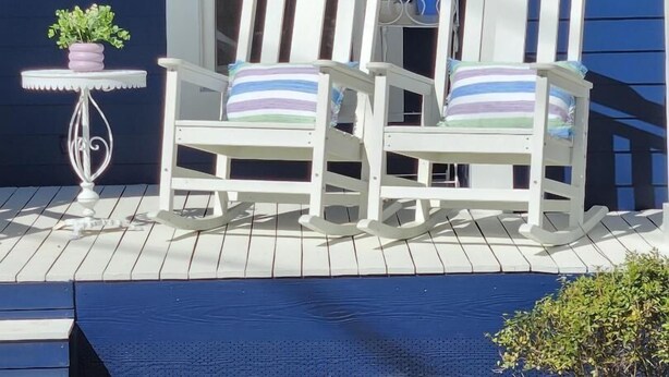 Une véranda avec deux fauteuils à bascule blancs, une petite table sur laquelle est posée une plante en pot et une jardinière suspendue.