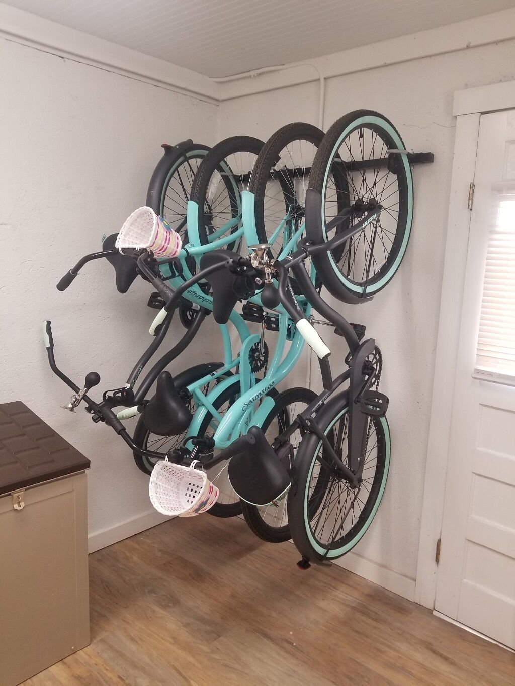 Una bicicleta color turquesa con una canasta y otra canasta rosa, montada en una pared de una habitación con suelo de madera y una puerta blanca.
