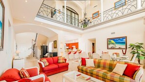 A spacious living area with a red sofa, a plaid sofa, and a glass coffee table. A balcony with a railing and a chandelier is visible above. - Two Story, 5 Bedroom Penthouse On Tamarindo Beach- New  (Tamarindo)