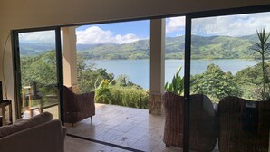 Habitación con grandes ventanales que ofrecen vistas a un lago y montañas. Hay sillones de mimbre y un sofá.