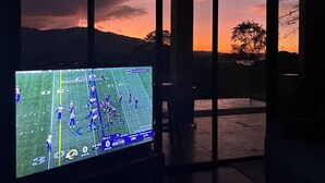 Un televisor mostrando un partido de fútbol, una ventana con vistas al atardecer y una zona de patio.