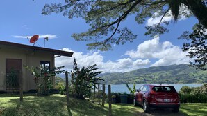 Un coche rojo estacionado frente a una casa con antena parabólica, rodeado de abundante vegetación y una vista panorámica de un lago.