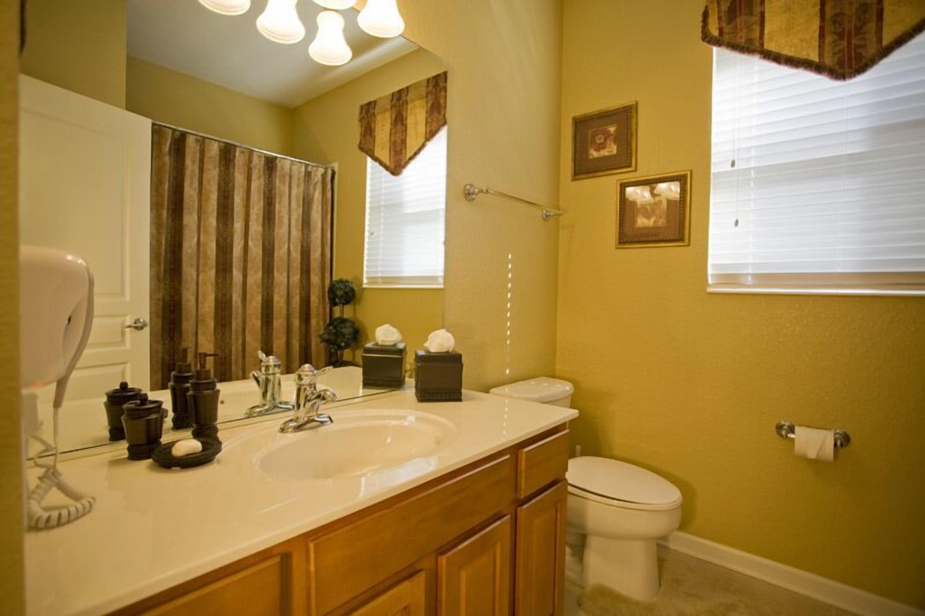 A bathroom with a white sink, vanity, and toilet. There are three framed pictures on the wall, a towel rack, and a window with blinds.
