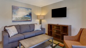 Een woonkamer met een grijze bank, een houten salontafel, een aan de muur bevestigde televisie en een schilderij boven de bank.