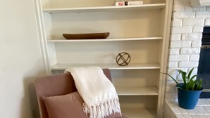Un coin douillet avec un fauteuil rose, un plaid blanc et une petite étagère ornée d’objets décoratifs.