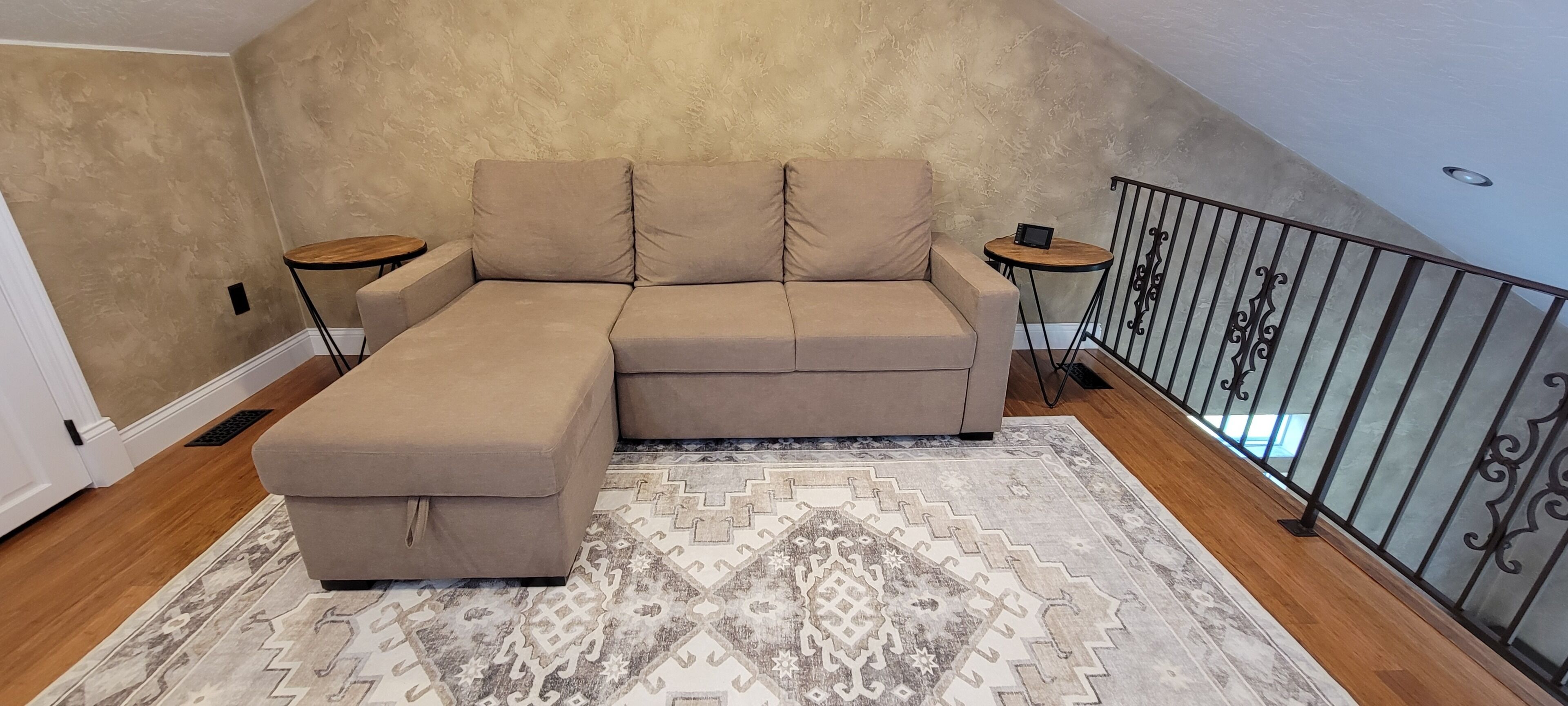 A beige sectional sofa with a matching ottoman, two small round tables, a patterned rug, and a balcony with a metal railing.